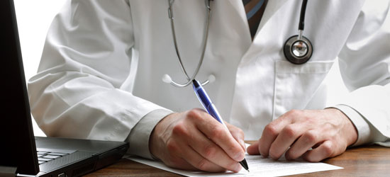 Crop of a doctor writing on a desktop with a laptop to the left